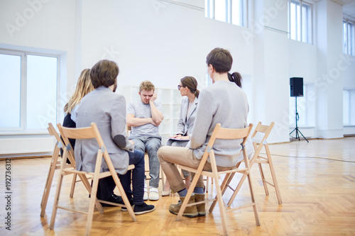 Group of young people with problems sitting in circle on chairs and looking at apathetic man during psychological session