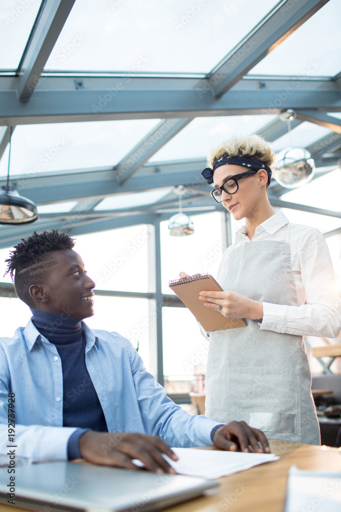 Young client sitting by table in cafe and ordering business lunch while waitress making notes