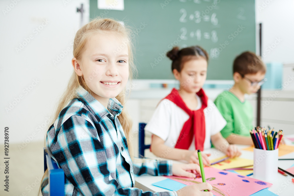 Fototapeta premium Happy blond schoolgirl smiling at you while sitting by desk and drawing at lesson