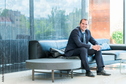 Portrait of a handsome mature businessman, relaxing on a couch in a business lounge, looking at the camera with a positive expression