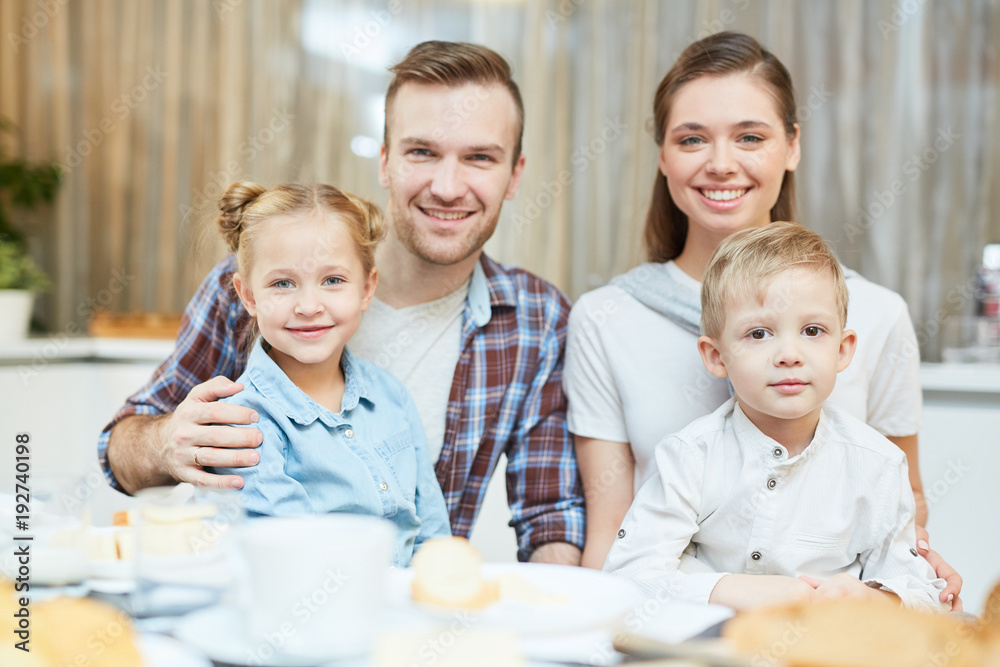 Happy young parents embracing their cute little son and daughter while sitting by served table Happy young parents embracing their cute little son and daughter while sitting by served table