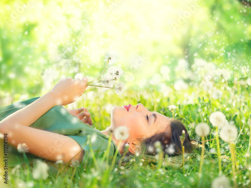 Beautiful young woman lying on green grass and blowing dandelions. Allergy free concept