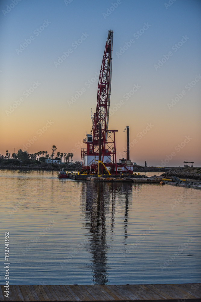 Long boom of marine crane extends into dawn sky as reflection in calm waters reaches toward the dock.