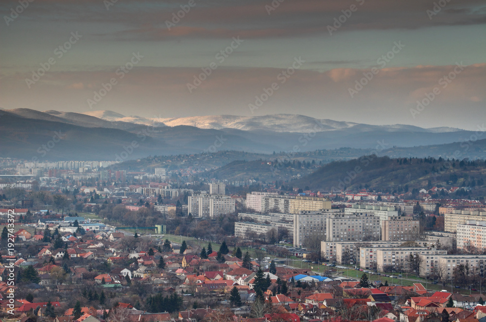 Elevated view of single-family houses and grey Eastern European socialist housing estates in Miskolc with wooded hills and snow-capped sunlit Bukk range of North Hungarian Mountains at winter Hungary