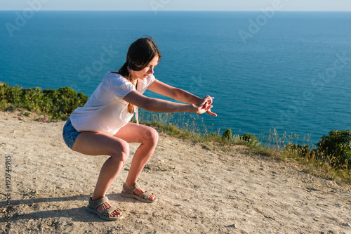 Woman doing sports on the mountain overlooking the sea, squats. Summer evening.