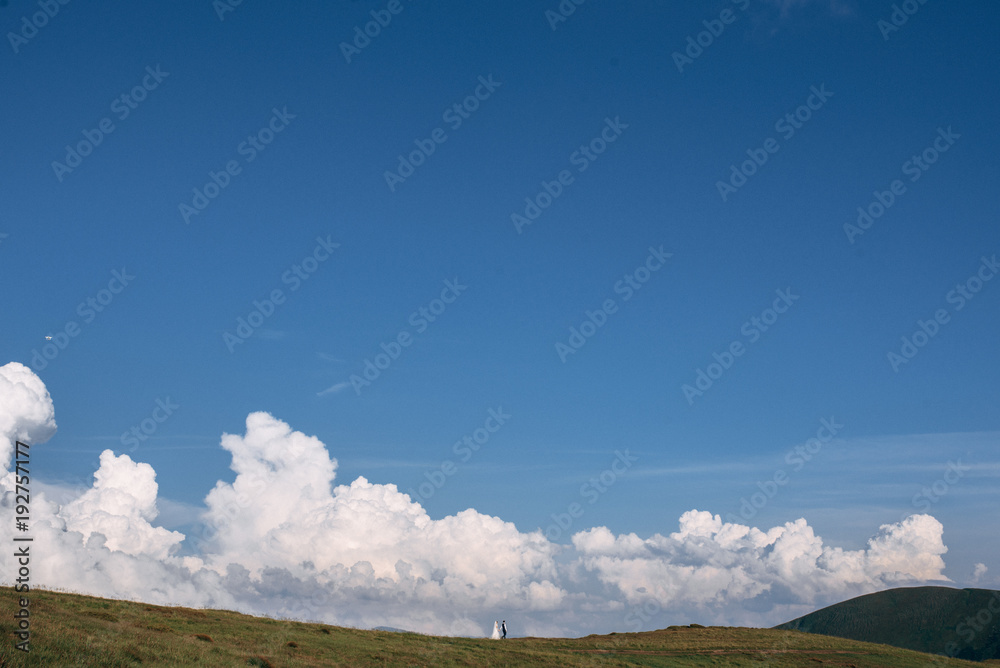 Bride and groom are walking on the green hills, sky with big clouds on background