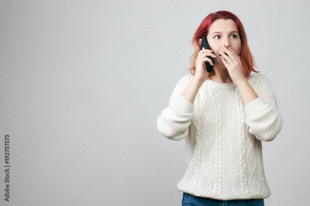 Shocked young woman in white sweater talking by smartphone with a frightened expression face over gray background with copy space.