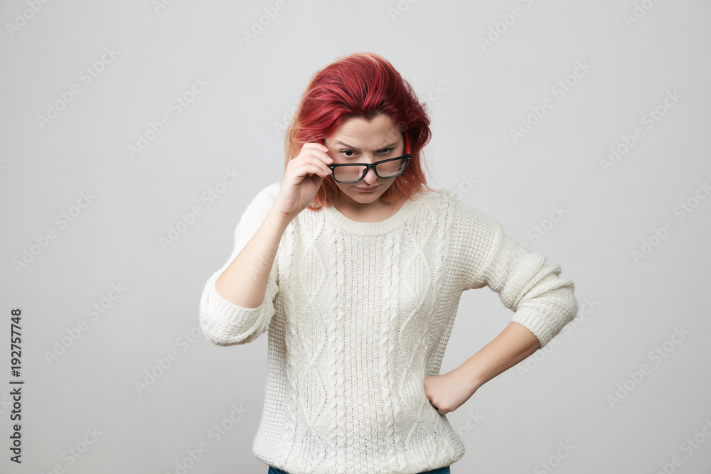Portrait of young woman in white sweater with confused and gloomy expression, correcting glasses and puts one hand on her hip, isolated over gray background. The girl is indignant Portrait of young woman in white sweater with confused and gloomy expression, correcting glasses and puts one hand on her hip, isolated over gray background. The girl is indignant