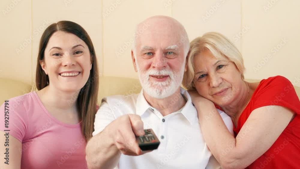 Senior couple and their teenage daughter sitting on couch and watching TV. Switching channels with remote control. Happy family enjoying time together Senior couple and their teenage daughter sitting on couch and watching TV. Switching channels with remote control. Happy family enjoying time together