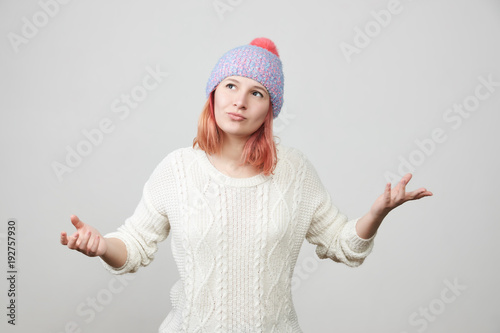 Young woman in knitted hat and sweater gesturing skeptical expressing disagreement with facial expressions over gray background with copy space. Indoor portrait. Reactions, attitude concept