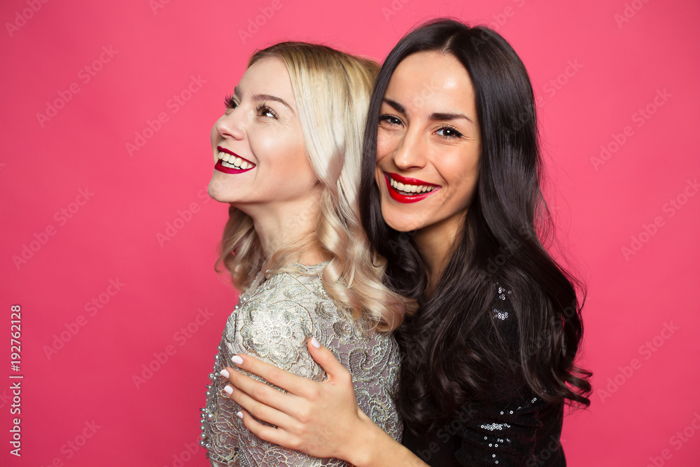 Obraz premium Friends forever. Close up photo of Two happy young beautiful smiling girlfriends in little black dresses posing and having fun on a pink background.