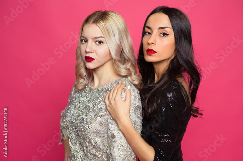 Fashion friends. Close up photo of Two stylish confident beautiful girlfriends in little black dresses posing on a pink background.