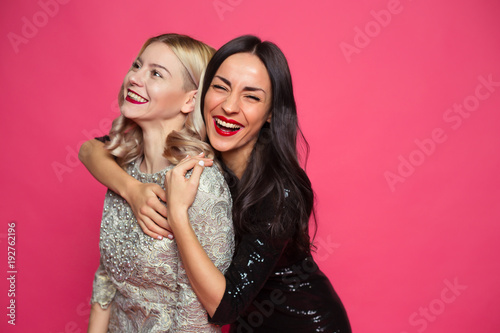 Friends forever. Close up photo of Two happy young beautiful smiling girlfriends in little black dresses posing and having fun on a pink background.