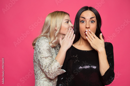 Gossip. Two young beautiful smiling girlfriends in little black dresses. The blonde tells a brunette story or gossip in her ear. photo