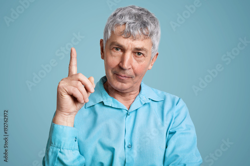 Experienced male pensioner in formal shirt raises fore finger as get brilliant idea for coming weekends, has confident look, isolated over light blue background. People, retirement and age concept