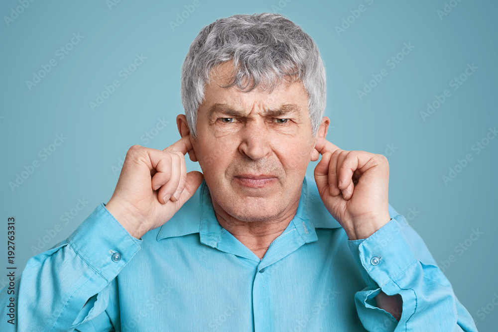 Portrait of upset annoyed mature man plugs ears with fingers, dressed in formal shirt, poses against blue background, doesn`t want to hear anything. Irritated elederly man ignores someone Portrait of upset annoyed mature man plugs ears with fingers, dressed in formal shirt, poses against blue background, doesn`t want to hear anything. Irritated elederly man ignores someone