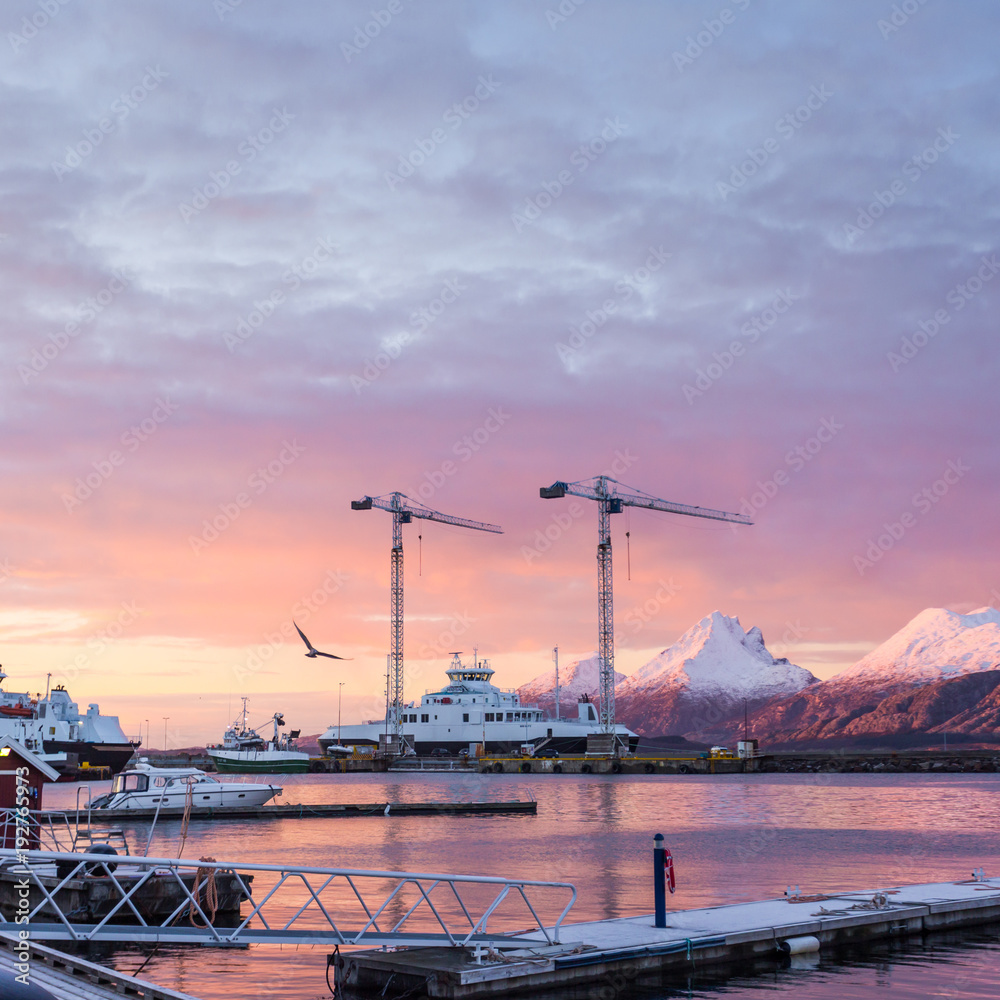 Beautiful sunrise in the port city Sandnessjoen. Berths, floating pontoons, tower cranes and moored ships on the background of the mountains and the pink sky reflected in the waters.