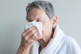 Head and shoulders view of older woman in white bathrobe blowing nose with tissue against neutral background (selective focus)