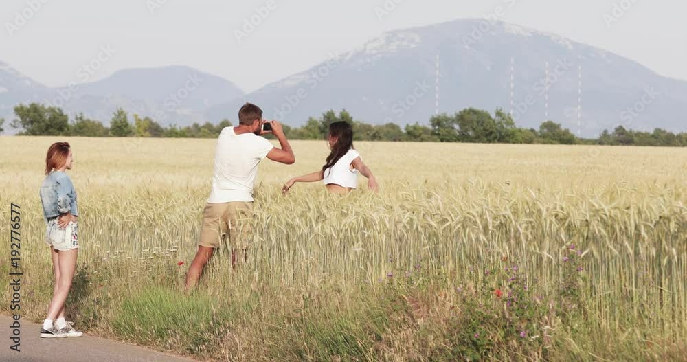 France, Provence, Two beautiful young girls and one man shooting photo in the field of wheat at sunset, happy girl with long hair, green trees on background France, Provence, Two beautiful young girls and one man shooting photo in the field of wheat at sunset, happy girl with long hair, green trees on background
