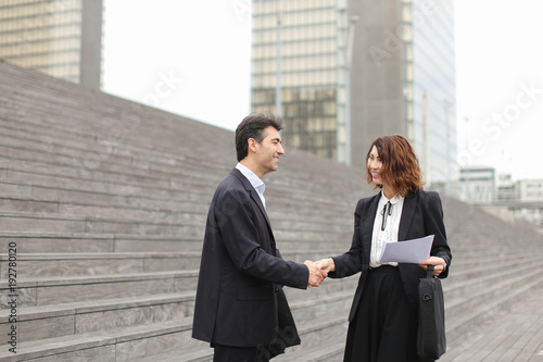 Layer and client standing on big gray stairs. Woman hold documents about court statement in hand. Man tell about very good end of litigation, and bouth smiling look really happy. Concept of pos photo