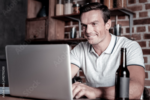 Time to take a break. Joyful millennial guy looking at a screen of his laptop with a cheerful smile on his face while sitting in his kitchen and relaxing with a bottle of beer.