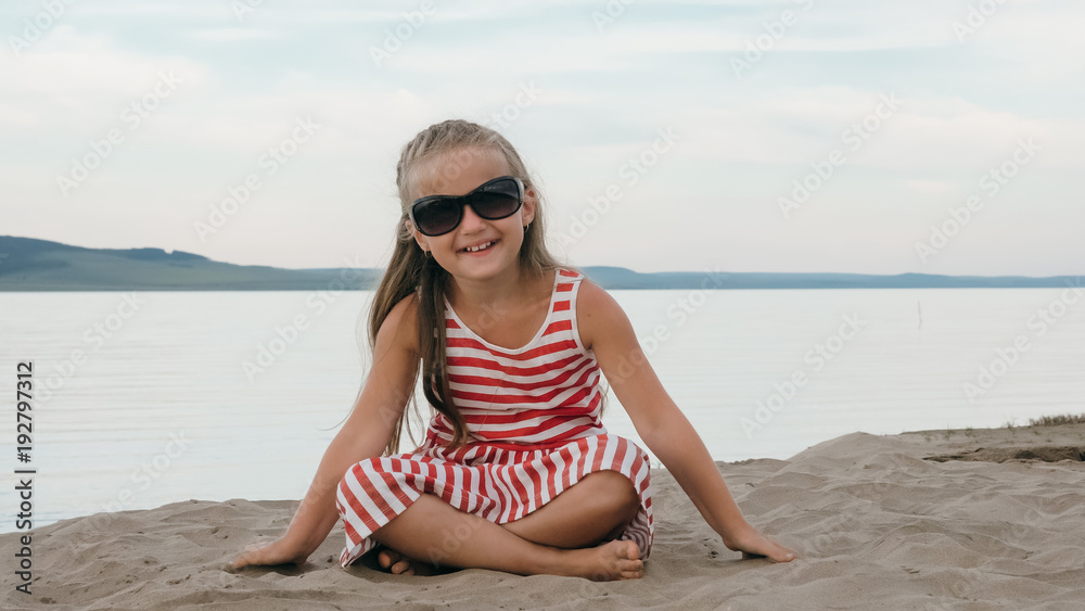 One beautiful teenage girl with brown hair outside on a beautiful summer day. The child is sitting on the beach. The sun was gone in the sunset. The kid is genuinely smiling and laughing.