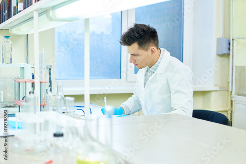 Profile view of handsome young researcher wearing rubber gloves and white coat sitting at desk and taking notes while carrying out experiment in modern lab