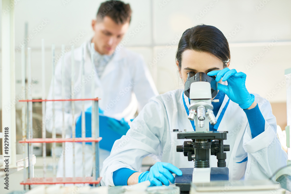 Confident dark-haired scientist wearing white coat and rubber gloves sitting at desk and using microscope, her male colleague standing behind and taking notes Confident dark-haired scientist wearing white coat and rubber gloves sitting at desk and using microscope, her male colleague standing behind and taking notes