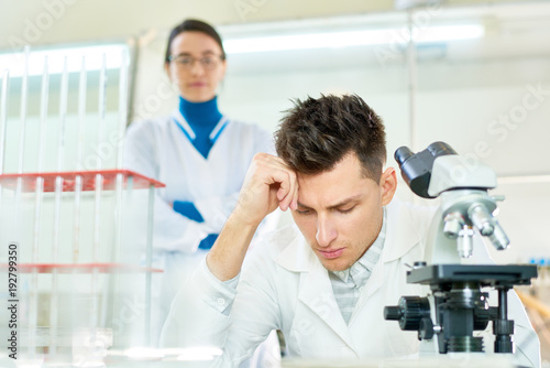 Concentrated young researcher wearing white coat leaning head on hand while sitting at desk  his pretty female colleague standing behind him  interior of modern lab on background