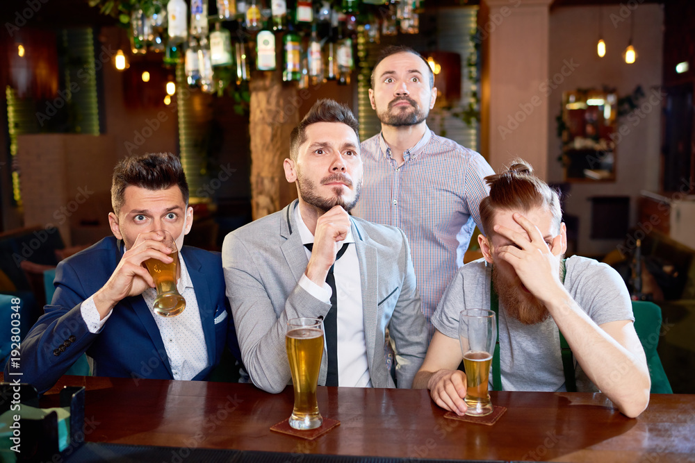 Nervous bearded friends gathered together in pub after work in order to watch football match and drink beer, one of them covering eyes with hand