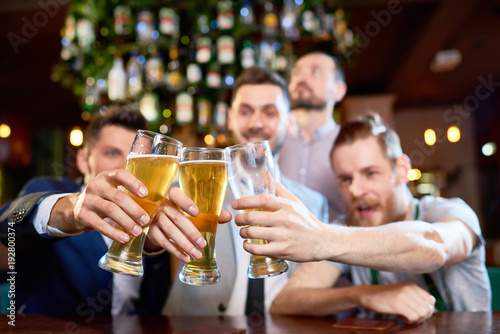 Group of cheerful friends clinking beer glasses together while sitting at bar counter and  celebrating end of work week  focus on foreground