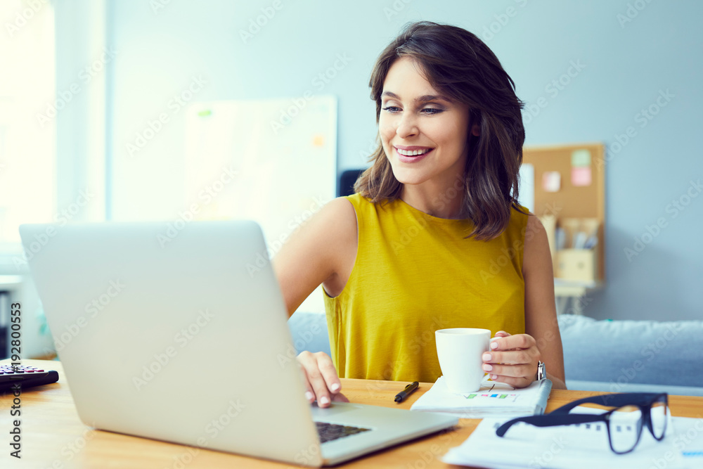 Attractive young businesswoman smiling while using laptop and drinking coffee in home office Attractive young businesswoman smiling while using laptop and drinking coffee in home office