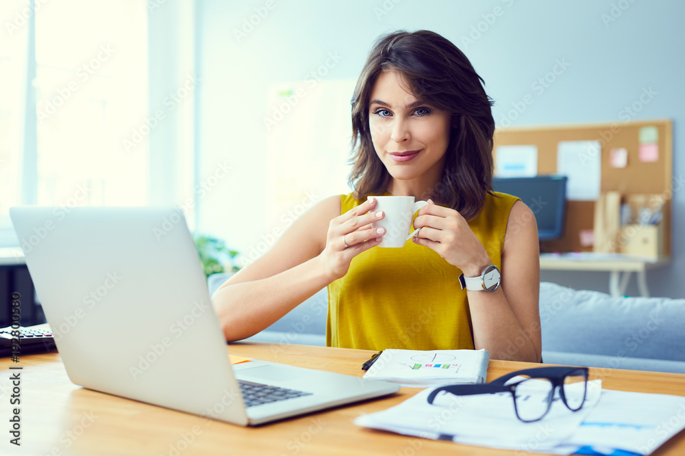 Beautiful young businesswoman smiling and looking at camera while holding coffee sitting at desk in office Beautiful young businesswoman smiling and looking at camera while holding coffee sitting at desk in office