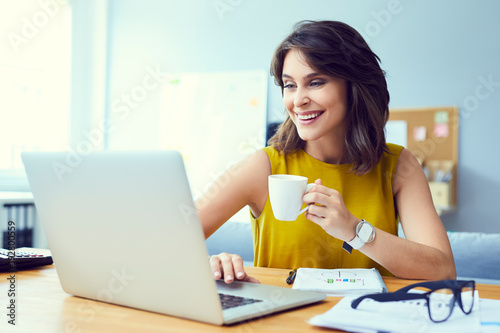 Gorgeous laughing young entrepreneur using laptop and drinking coffee at her desk in modern home office