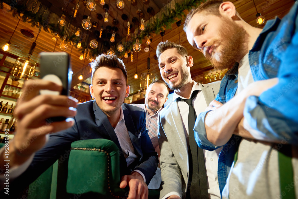 Obraz premium Smiling bearded friends taking selfie with help of smartphone while having long-awaited gathering in modern pub