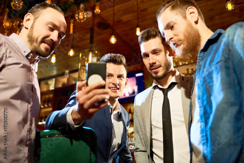 Low angle view of cheerful friends looking through funny photos on smartphone while spending evening in modern pub