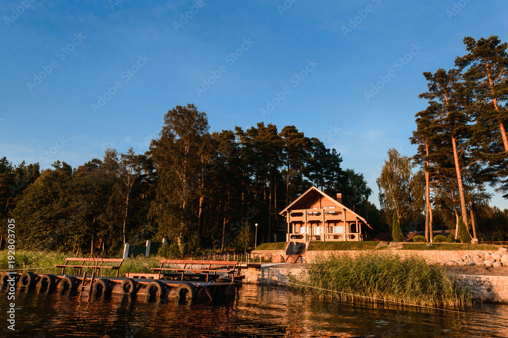 Obraz premium Modern russian village in national russian style on the lake coast. Good place for camping, fishing, hunting and relax. Summer sunset sky reflection in calm warm water.