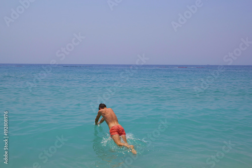Young guy jumps into the blue water with sea foam. Turquoise water with white pebbles on the beach. Sea line meets the horizon making beautiful panoramic view. Handsome man dive in pink swim trunks. © Vadym