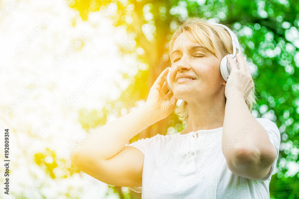Happy elderly woman listening music enjoying headphone in the park, relaxing time concept in Morning sun light