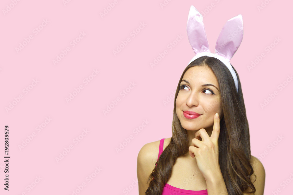 Attractive young woman with bunny ears looking to the side thinking or desire your product isolated on pink background. Copy space.