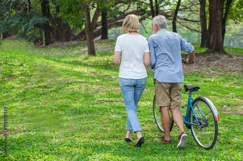 Senior couple in love walking with bicycle in park backside