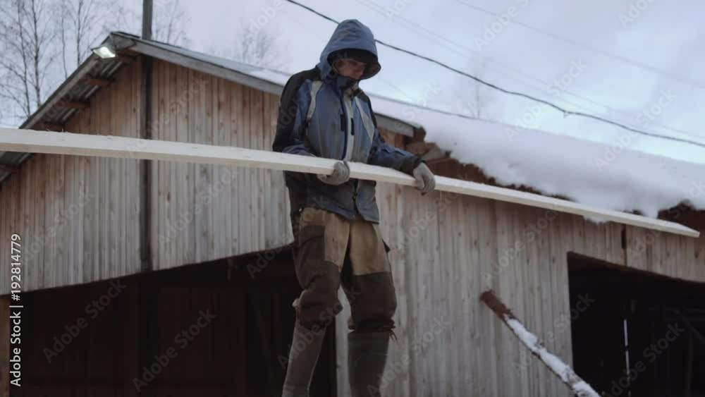 Caucasian man porter in dirty clothes carrying wood plank at sawmill factory yard on cold winter day Caucasian man porter in dirty clothes carrying wood plank at sawmill factory yard on cold winter day