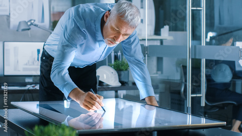 Senior Architectural Engineer Draws Building Concept on a Graphics Tablet Display Vertical Touchscreen Table. Clean Minimalistic Office, Concrete Walls Covered by Blueprints.