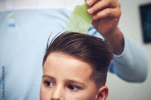 Barber is making a hairstyle to Caucasian boy in barbershop.