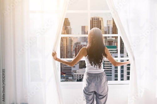 Woman waking up and opening window curtains at home