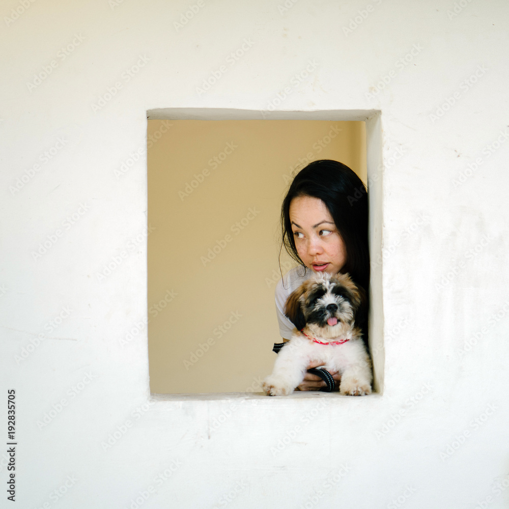 A young Pan Asian woman is carrying her dog and peeking through a window and smiling in a candid, relaxed and authentic manner. A young Pan Asian woman is carrying her dog and peeking through a window and smiling in a candid, relaxed and authentic manner.