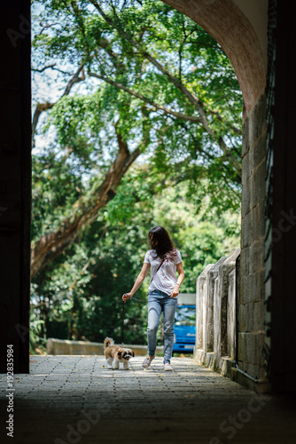 An image of a beautiful and young Asian woman with her shih tzuh dog walking in the park. She is smiling while enjoying her day under the sun and as they play along.