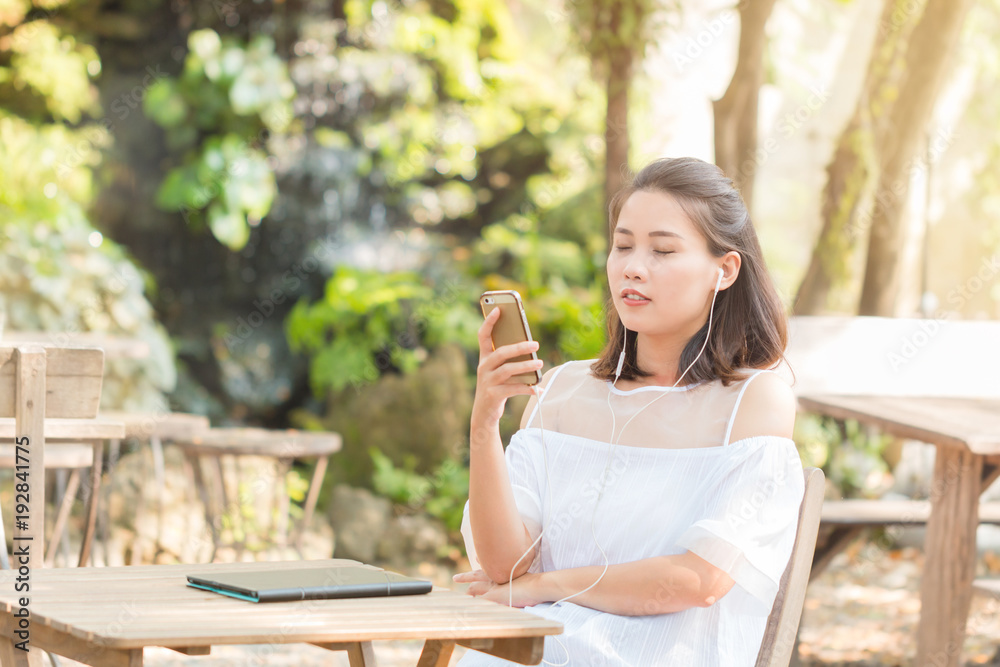 Portrait of Attractive young Asian woman wearing white casual relaxing and listening to music on smartphone at the coffee shop park outdoors.