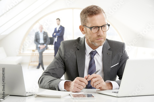 Businessman using laptop at the office. Portrait of senior financial male director wearing suit and tie sitting at desk and working. Uncrecognizable business people standing at the background. 