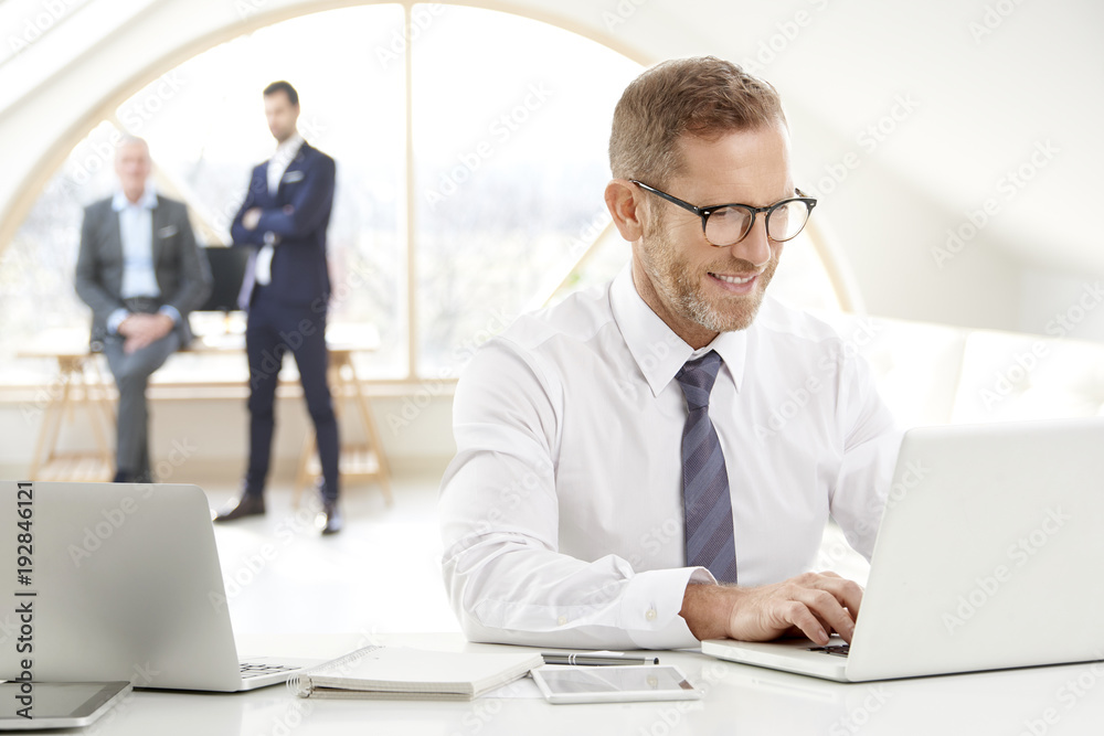 Businessman using laptop at the office. Portrait of senior financial male director wearing suit and tie sitting at desk and working. Uncrecognizable business people standing at the background. Businessman using laptop at the office. Portrait of senior financial male director wearing suit and tie sitting at desk and working. Uncrecognizable business people standing at the background.
